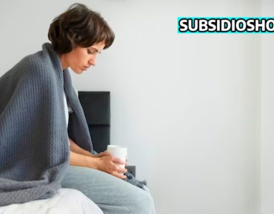 mujer sentada en la orilla de la cama enferma con un vaso en mano, arrba a la derecha el loo de subsidiohoy.com